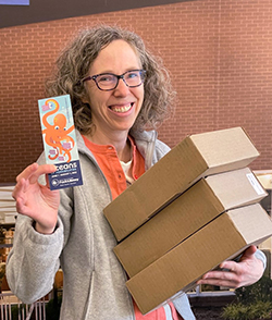 Graphic design coordinator Sue (woman with curly hair and glasses) holds boxes full of Summer Reading bookmarks